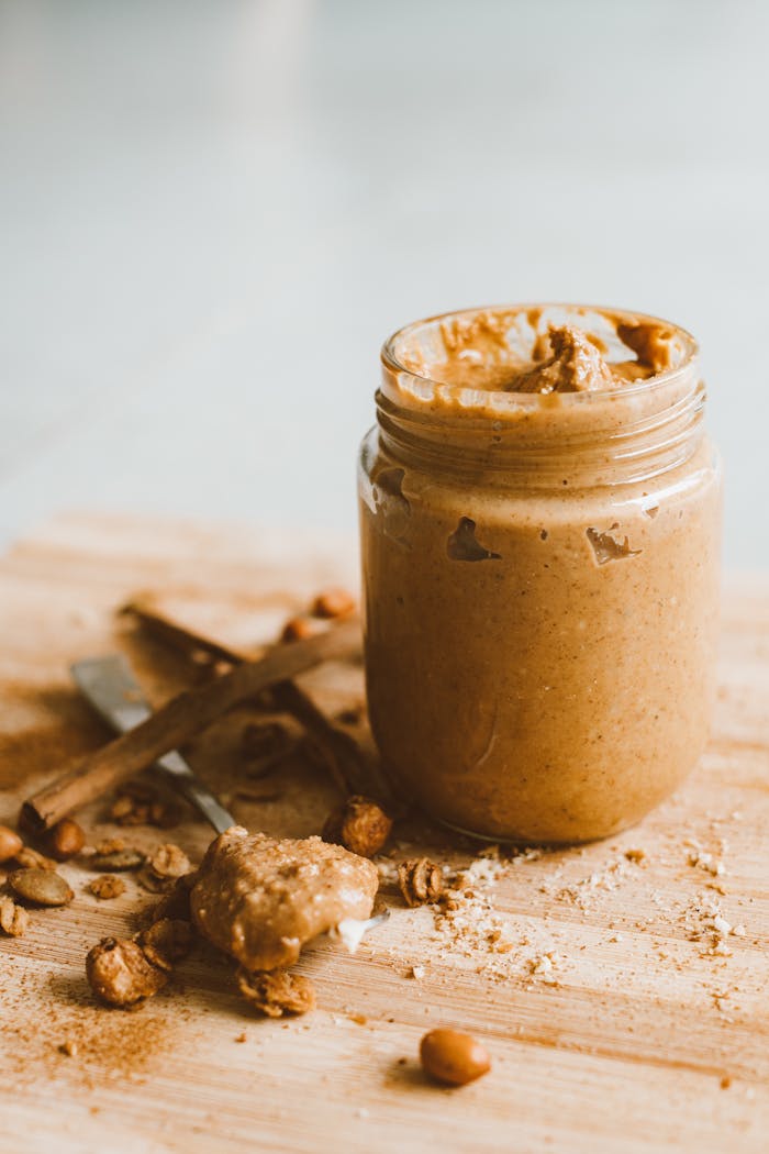 A jar of creamy peanut butter with scattered peanuts and spoon on a wooden surface.